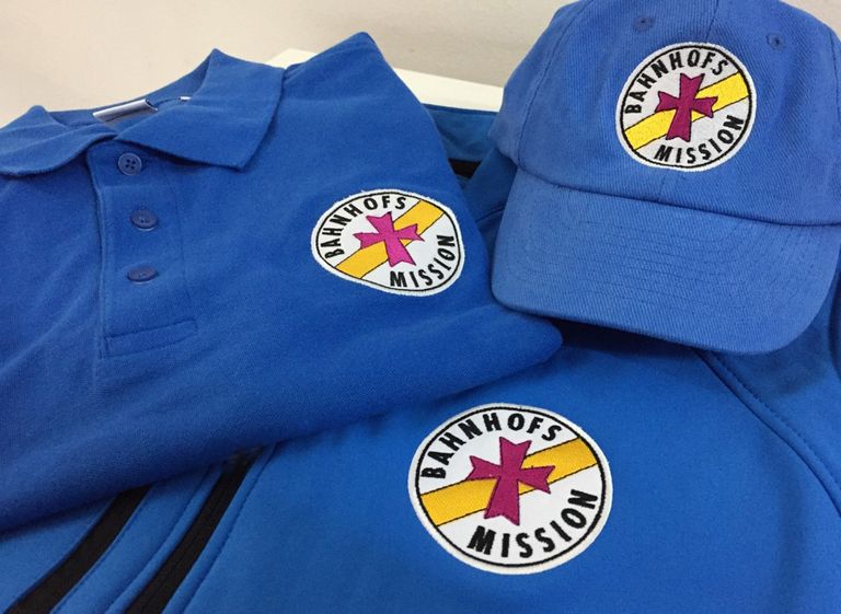 Poloshirt, Basecap und Jacke mit Stickereien der Bahnhofs Mission